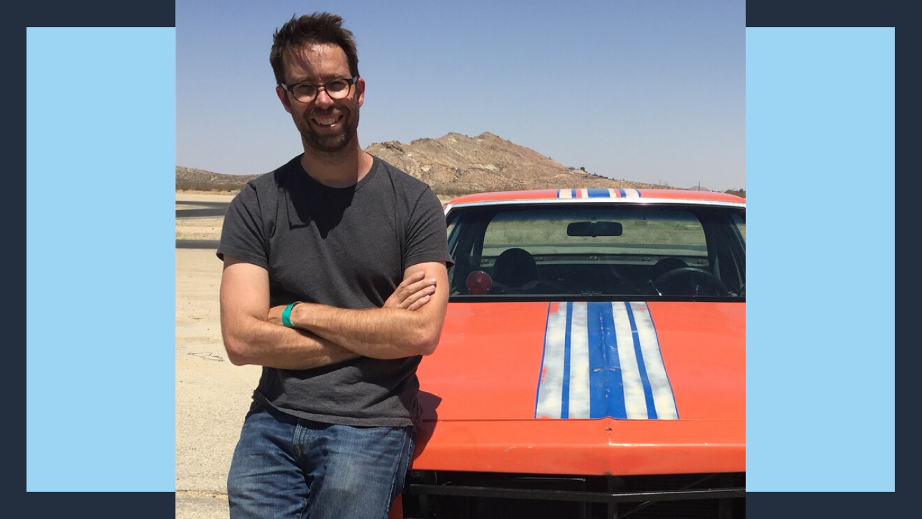 Jon crosses his arms and leans against the hood of a vintage muscle car.