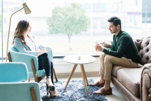 A man sits on a couch across from a woman therapist who is sitting in a blue chair and writing on a notepad. 