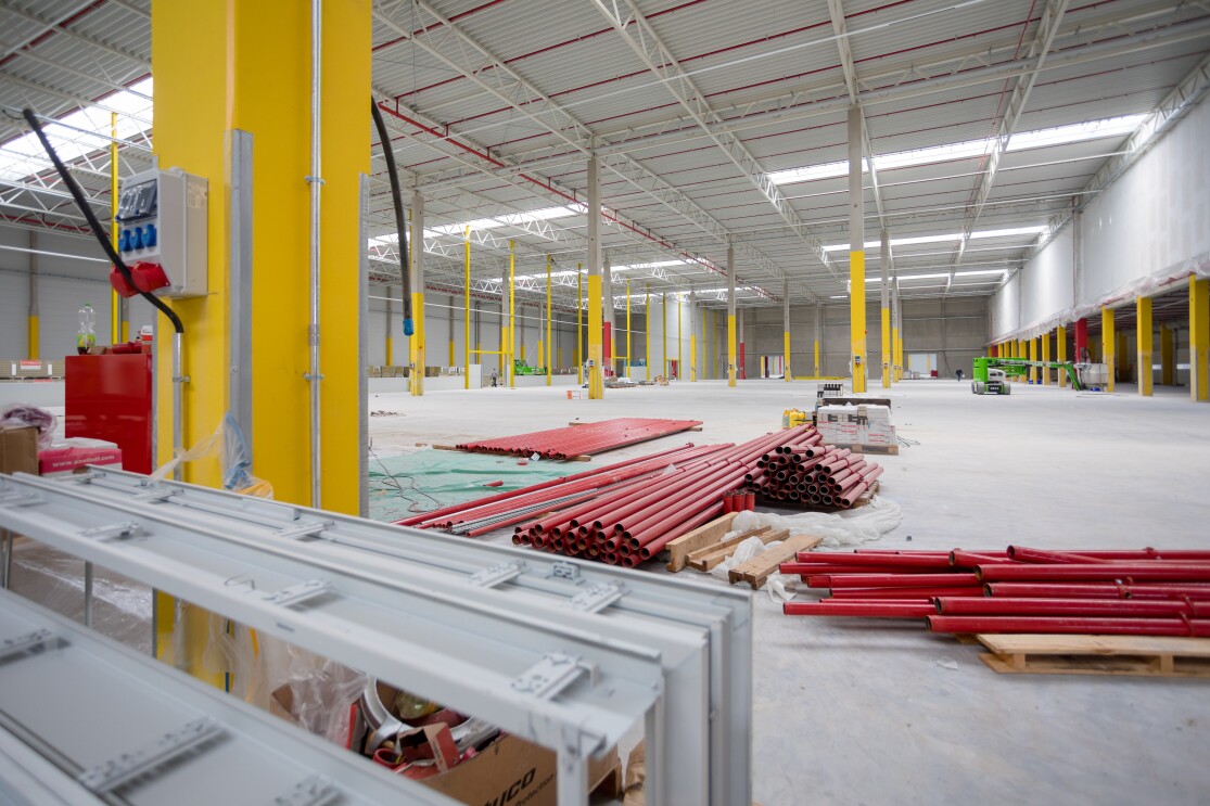 Eine leere weiße Halle mit gelben Stützpfeilern. Auf dem Boden sind zahlreiche rote Rohre gestapelt.