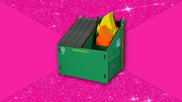 Miniature toy dumpster fire desk caddy holding bright flame notecards, set on vibrant pink glitter surface.