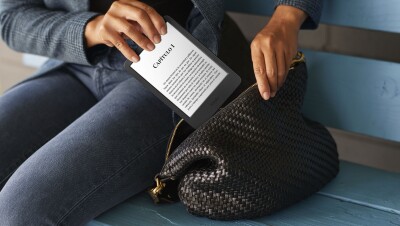 En un banco de madera azul, está sentada una mujer que guarda su Kindle en un bolso. El plano es corto y enseña manos y piernas de una mujer. La mujer va vestida con unos jeans u una americana de cuadros escoceses. La mujer lleva un bolso de piel de color negro. El Kindle que está guardando se ve el texto Capítulo 1 y un texto que no se puede leer.