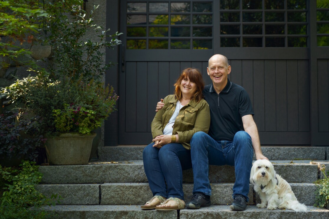 A woman and a man site with a dog on stone steps. They are smiling into the camera. She is wearing blue jeans and a green shirt. He is wearing blue jeans and blue shirt. 