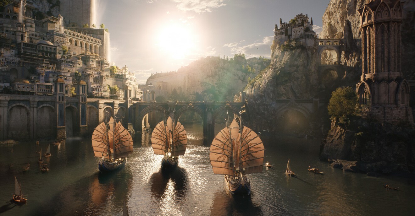 A scene from the Lord of the Rings: The Rings of Power. Ships with circular sails move through a bay. There is a bridge and a city on the hill surrounding the bay.