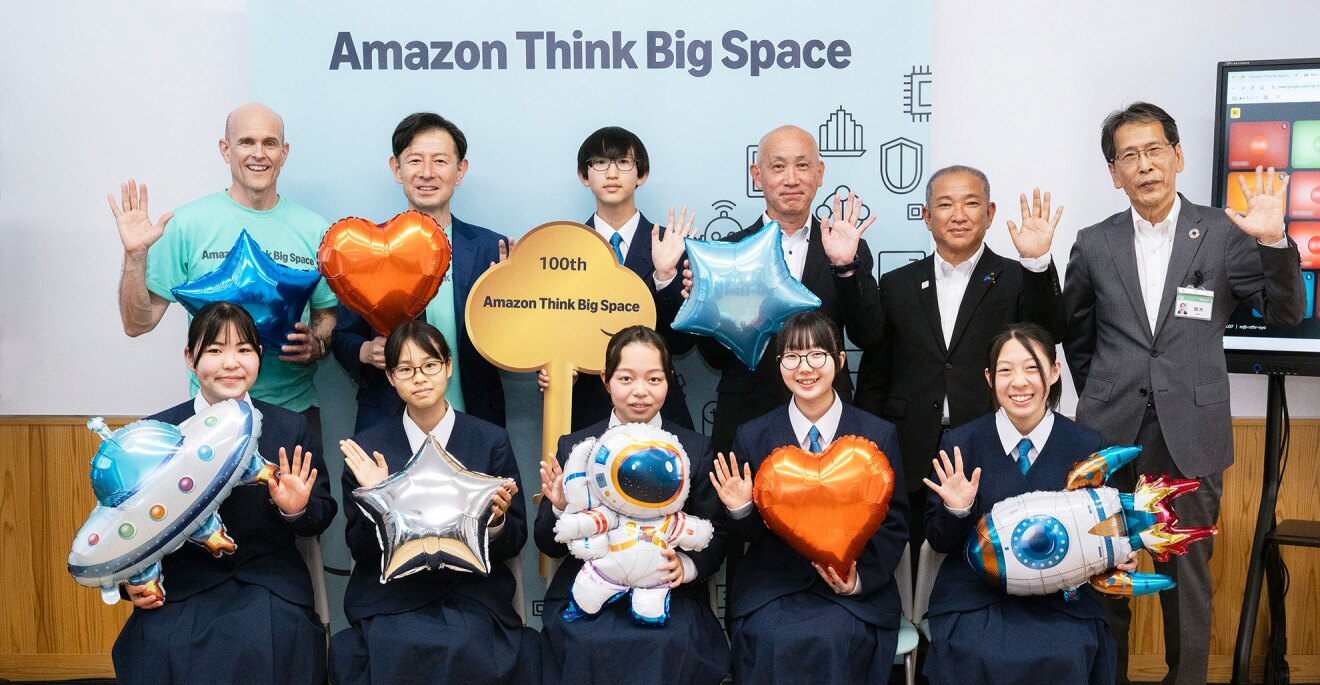 Amazon Think Big Spaceと書かれた背景の前に並び、手を振る人々。前列は制服の生徒5人が座り、後列にはスーツやTシャツ姿の成人5人と、中央に「100th」と書かれた金色の鍵型オブジェクトを持った生徒が立っている。他の8人は宇宙船や宇宙飛行士、ロケットを模したバルーンを持っている。