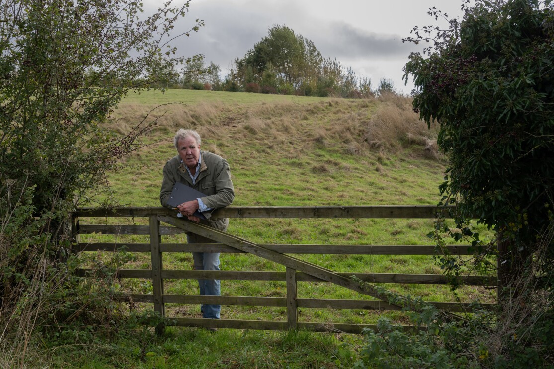 An elderly man holding a clipboard and leaning against a fence with a farm in the background