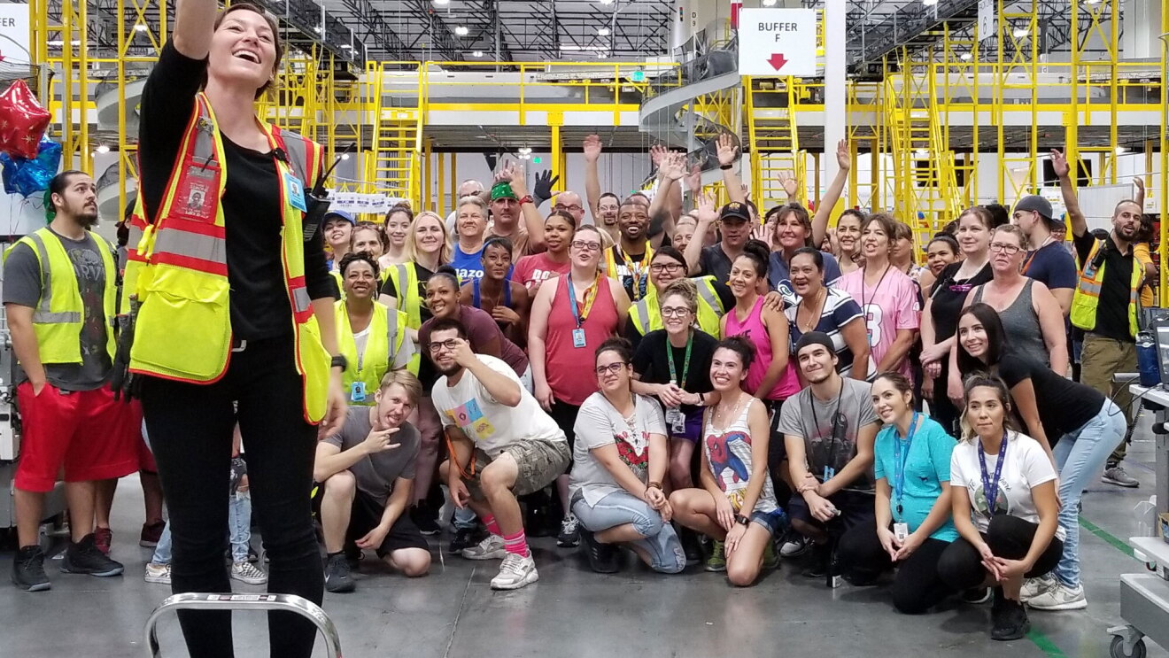 An Amazon associate stands on a step stool, with her arm extended, holding her iPhone in order to capture a selfie. Behind her, dozens of Amazon associates pose for the photo.