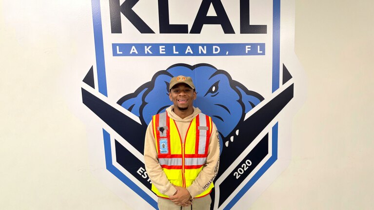 An Amazon employee wears a yellow and orange safety vest and smiles while standing in front of KLAL logo.