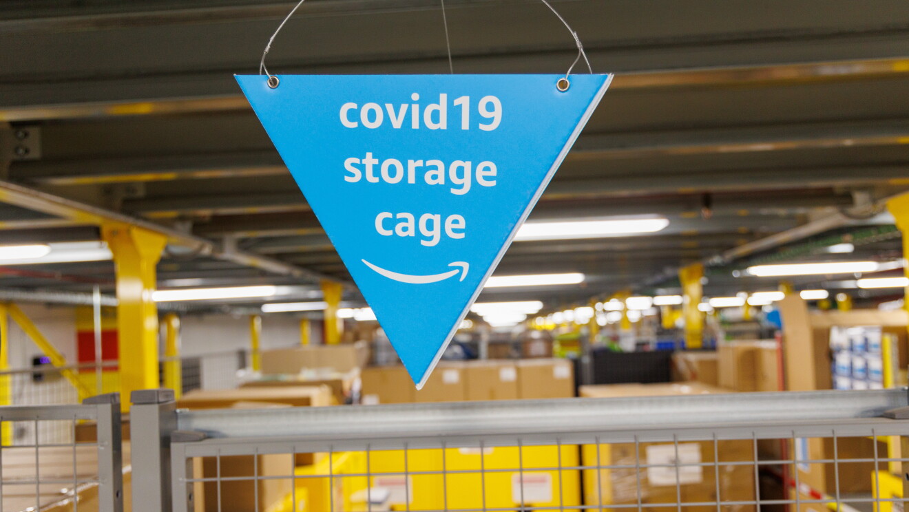 Dentro de un centro logístico, en primer plano hay una banderilla de cartón de color azul con este mensaje: COVID19 storage cage. Está encima de una valla de color gris. De fondo cajas de cartón y de plástico de color amarillo.  