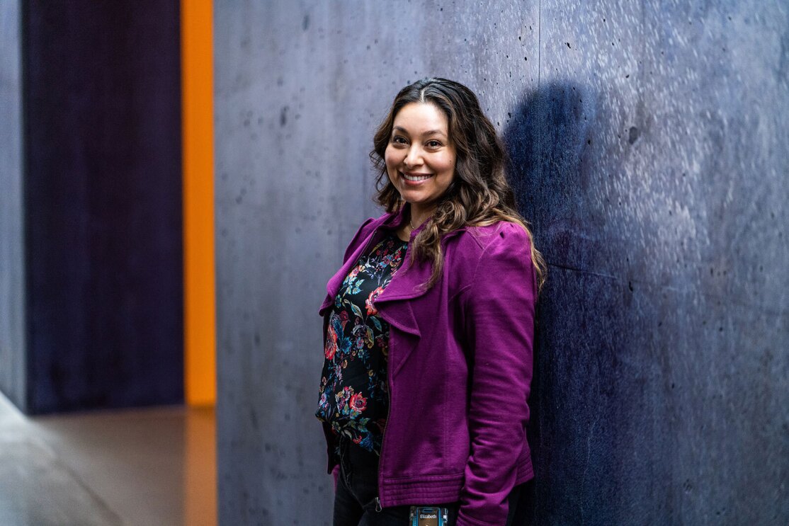 An image of a woman smiling for a photo standing in front of a dark gray and blue wall at an Amazon office building.