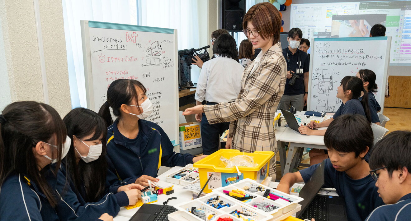 教室でジャージや体操着姿の生徒たちが、班に分かれて組み立て作業に取り組む。机の上にはノートPC、黄色い収納ボックス、各種パーツが入った白いトレー。チェック柄の上着を着用した人物が立って話をしている。奥のホワイトボードには図や文字の書き込み。さらに奥にも別な班の机、ホワイトボードがある。