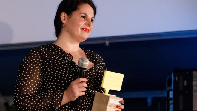 Julia Ritte hat den Preis steht auf der Bühne. In einer hand hält sie ein Mikrofon, in der anderen den UdZ-Pokal. Sie blickt freundig und gerührt ins Publikum.