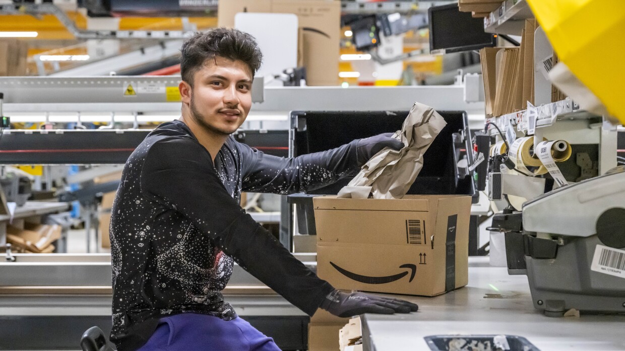 Trabajador de centro logístico sentado en una estación preaparando una caja. Lleva el pelo corto, pero la parte de arriba más larga. Lleva camiseta negra con un planeta en la parte delantera. Lleva guantes de protección y unos pantalones azules.