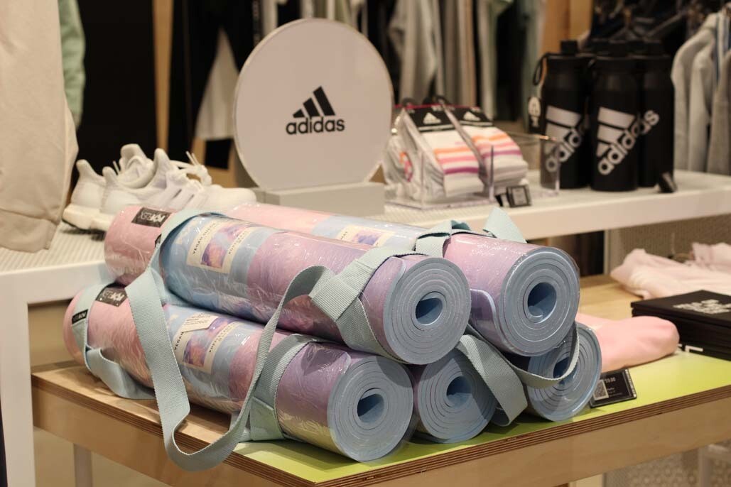 A stack of products including yoga mats and athletic gear sit atop a shelf in a store with an Adiddas sign next to them.