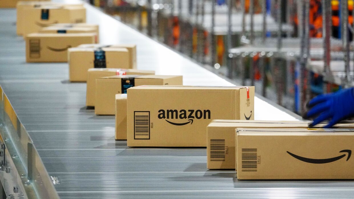銀色のコンベア上を、Amazonロゴ入りの段ボール箱がたくさん流れている。最も手前の箱の上に、青い手袋を付けた手が見える