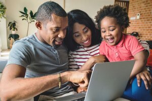 A family of three smile as they sit right next to each other on a couch looking at a laptop.