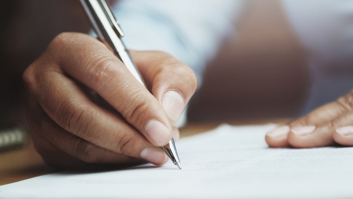 Primer plano de una mano con un bolígrafo firmando un documento. La otra mano está apoyada encima del documento. Se ve una camisa blanca pero desenfocada. El bolígrafo es plateado.