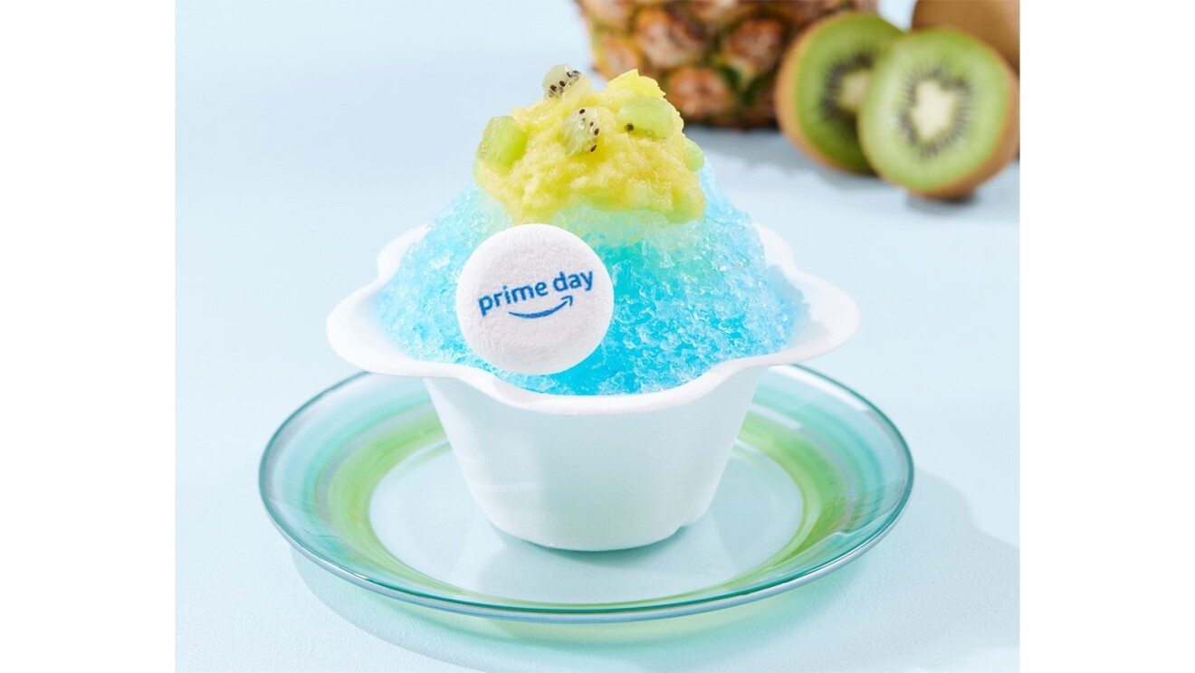 青いかき氷がテーブルの上に置かれている。そのかき氷には黄色く切った果物と白い円形のPrime Dayロゴがトッピングされている。後ろにはキウイとパイナップルが見える。