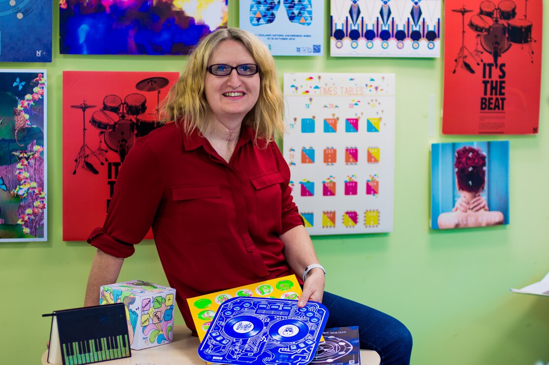 A blonde woman wearing glasses, jeans, and red button up shirt is seated on a tabletop in a room painted green, with myriad artwork hung on the wall.