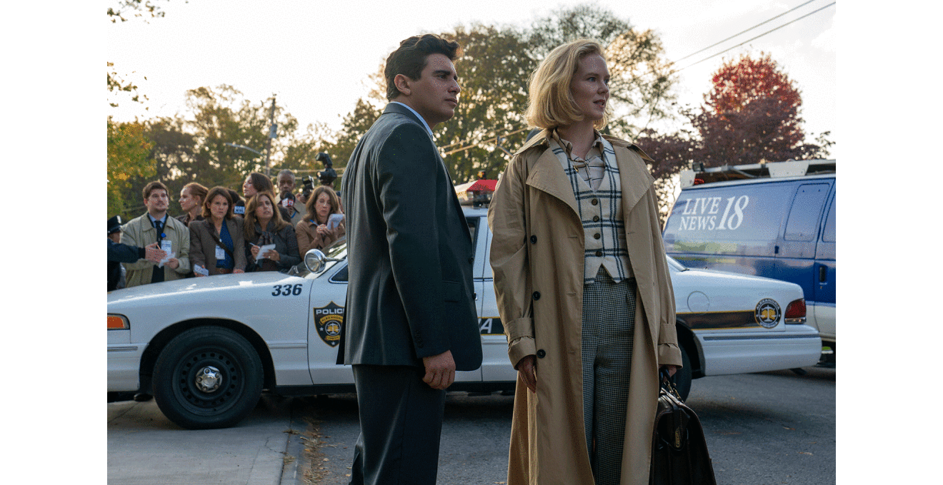 規制テープが張られた屋外で、スーツ姿の人物とトレンチコートの人物が立っている。2人の背後にパトカーと報道車両、手にメモを持った報道陣。