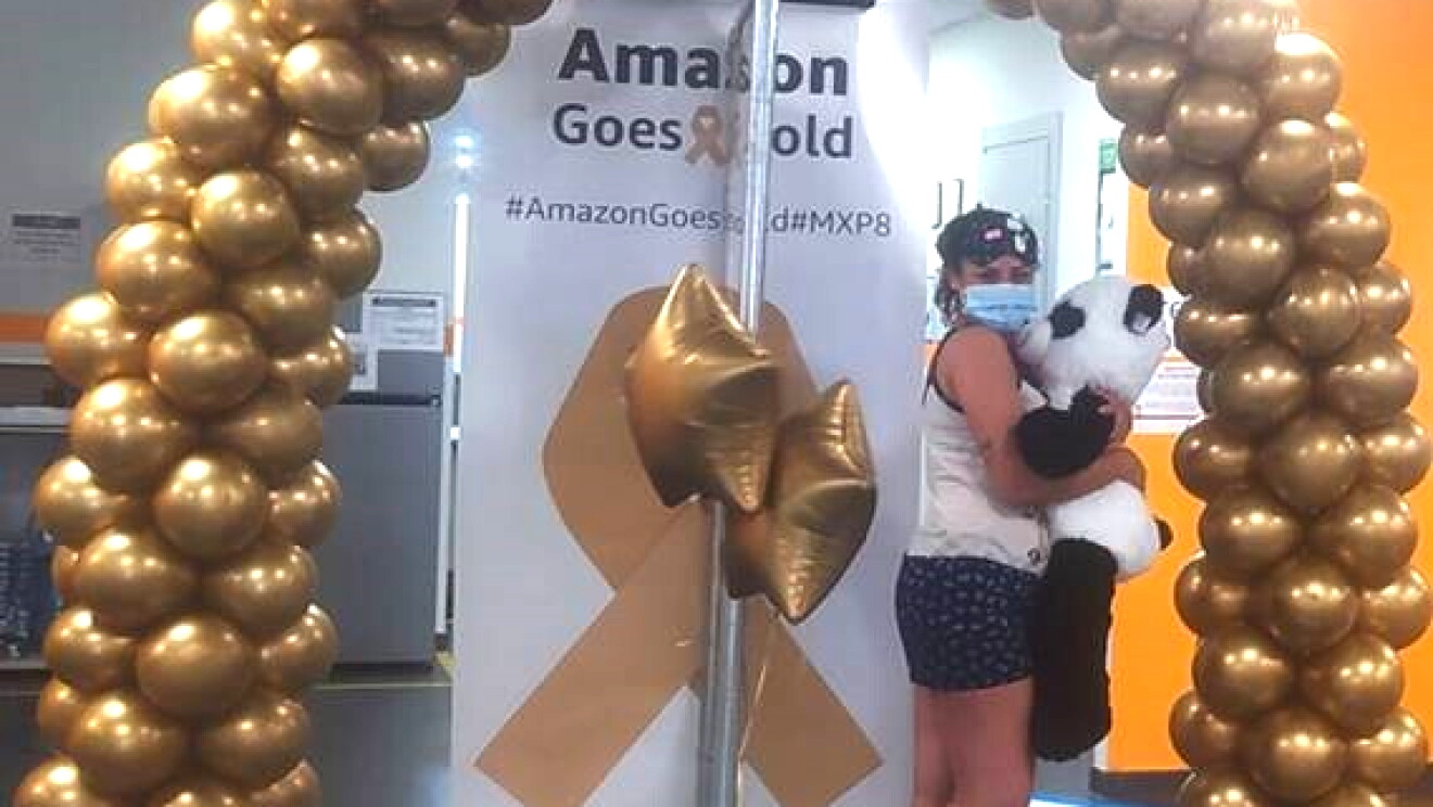 Dipendente Amazon posa con un peluche a forma di panda sotto un arco di palloncini dorati e accanto un cartellone con la scritta Amazon Goes Gold 2021