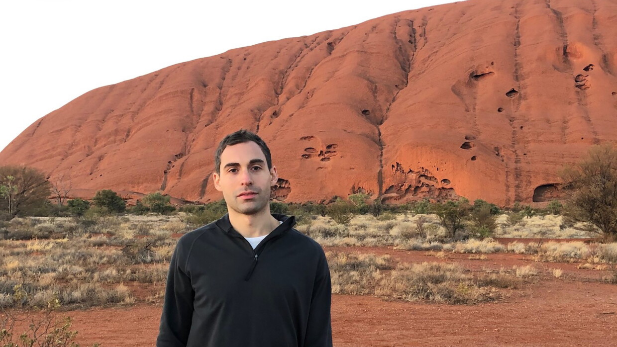 A man wearing a half-zip jacket and t-shirt stands in an arid location near the brush