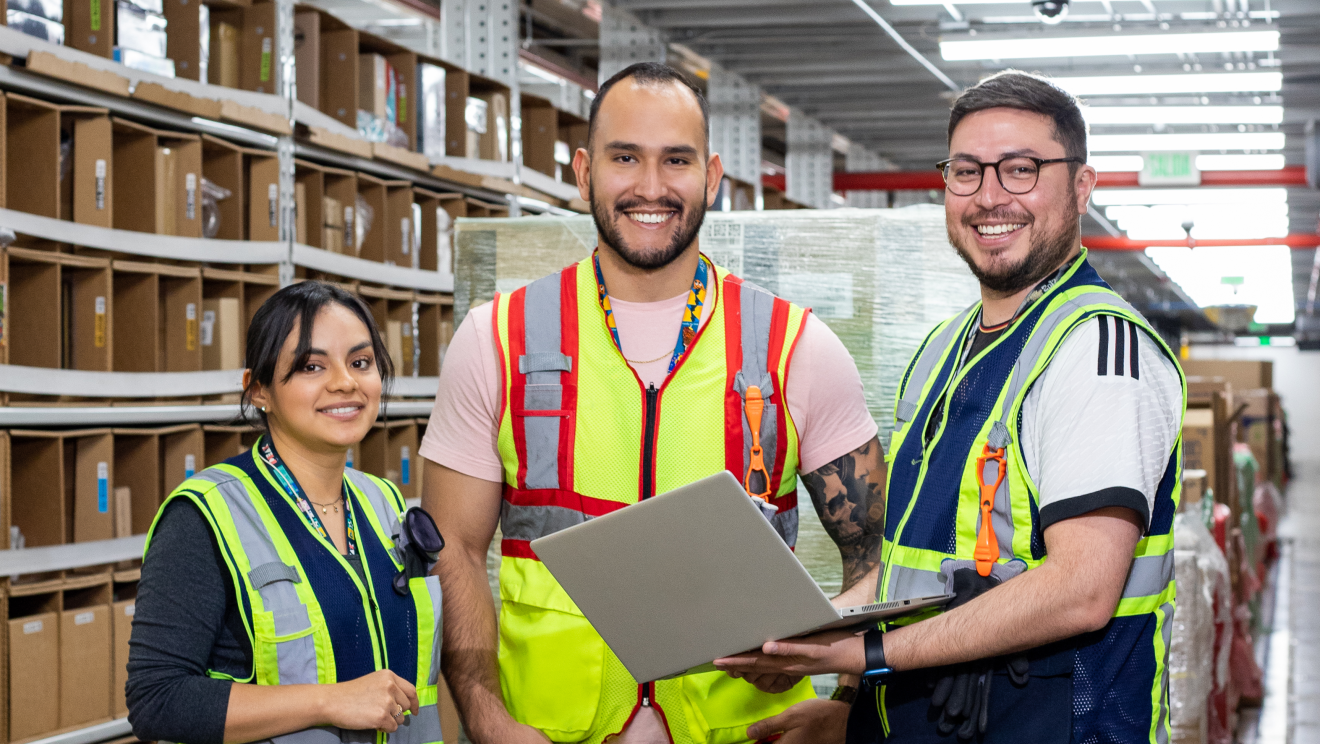 Tres trabajdores de un centro logústico depie, en un pasillo mirando a cámara. 