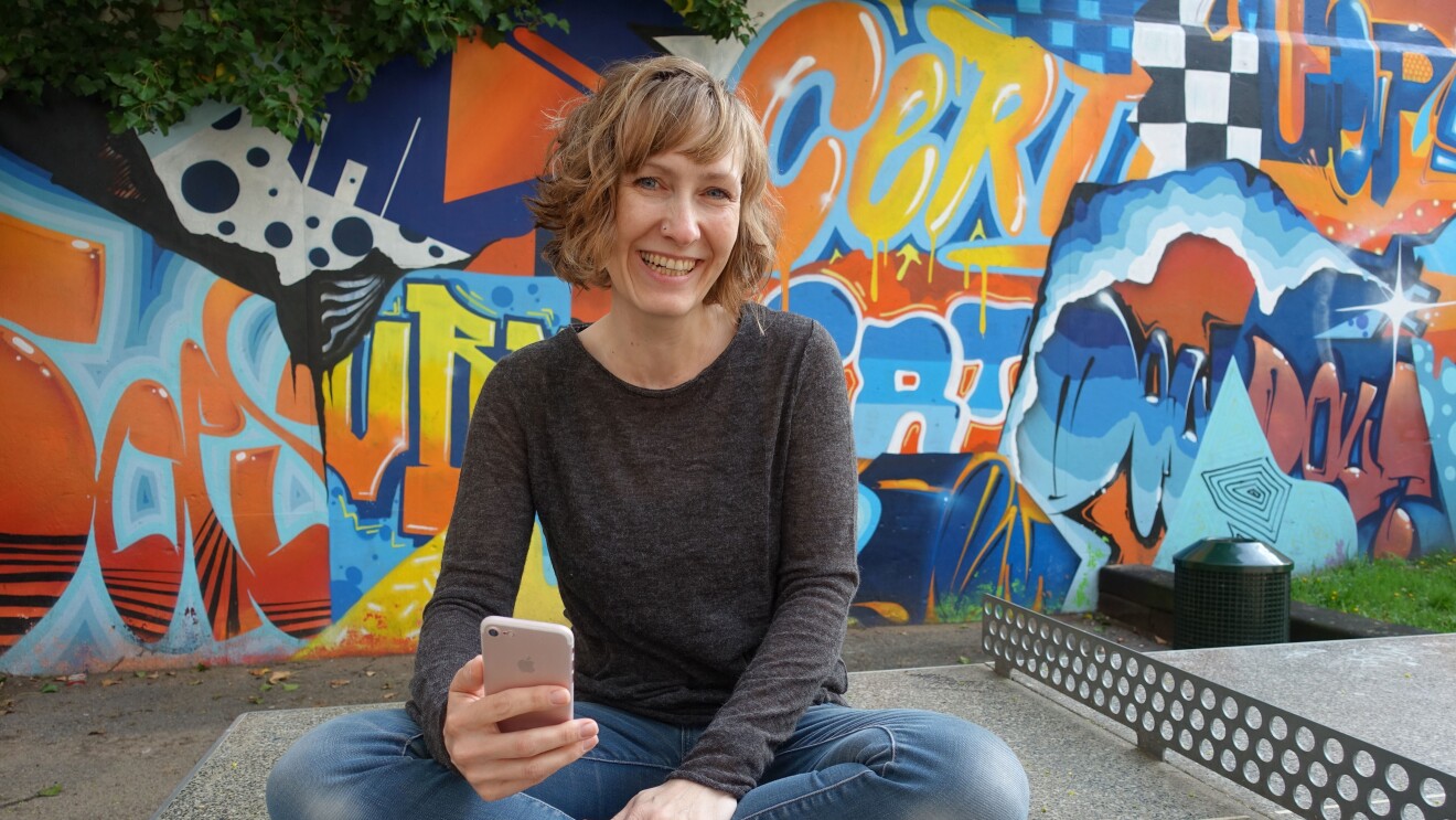 Eine Frau mit kurzen blonden Haaren und einem grauen Pullover sitzt auf einer Tischtennisplatte und lacht in die Kamera.