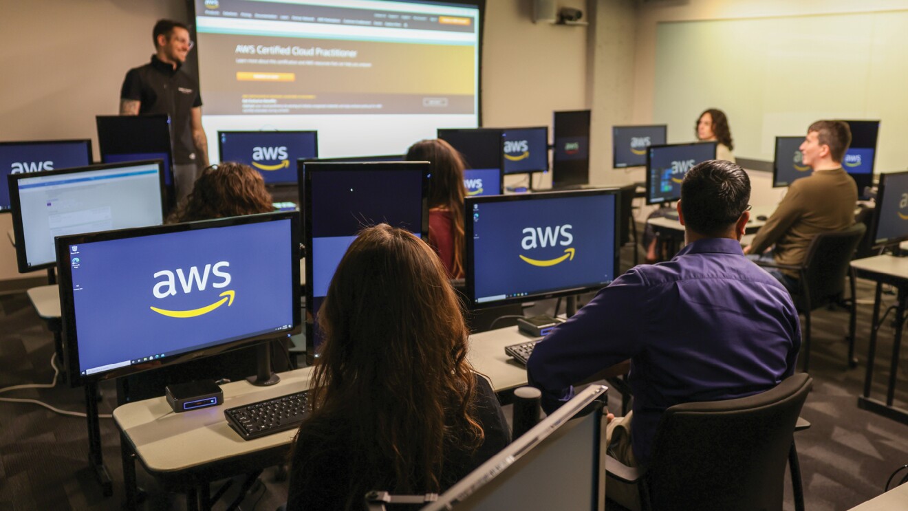 A man stands in front of a classroom, a projector screen next to him has text that says "AWS Certified Cloud Practioner" on it. In front of him, students sit at computers and look in his direction.