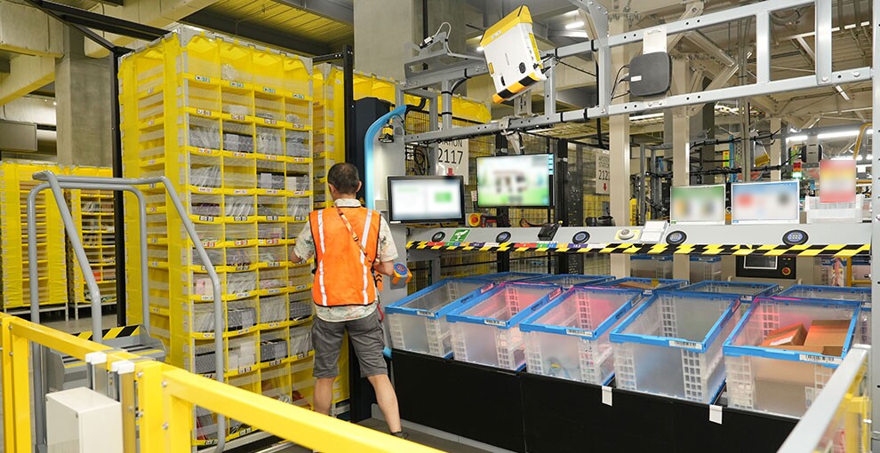 物流施設の作業ステーションで立って作業するオレンジ色ベストを着用した人物の後ろ姿。左側に背が高く細かく区分けされた黄色い棚、右側に半透明の収納ボックスが並び、目の高さにはモニター2面。