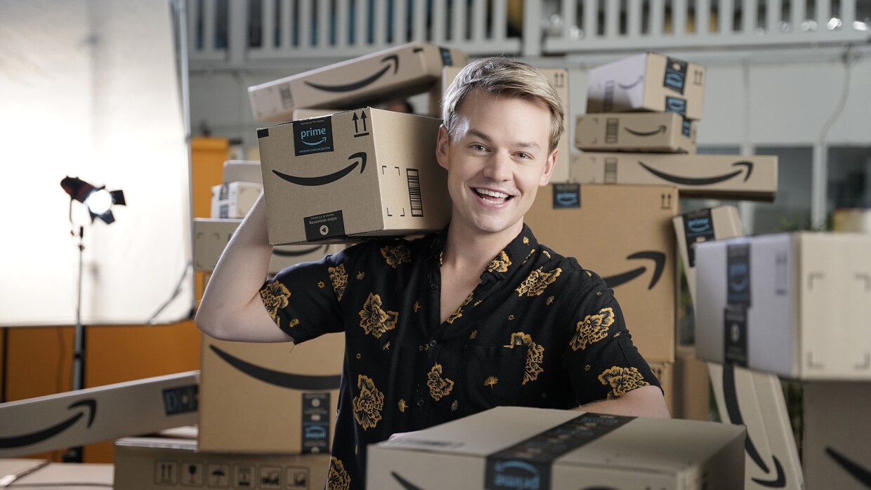 A young man wearing a button down shirt smiles at the camera, while holding a few Amazon boxes. Behind him is another, tall stack of Amazon boxes.