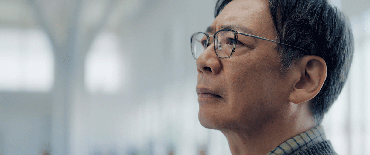 白と灰色のぼやけた背景、右半分に中年男性のクローズアップ。眼鏡をかけ、少し斜め上を見上げている。眉間にしわをよせている。