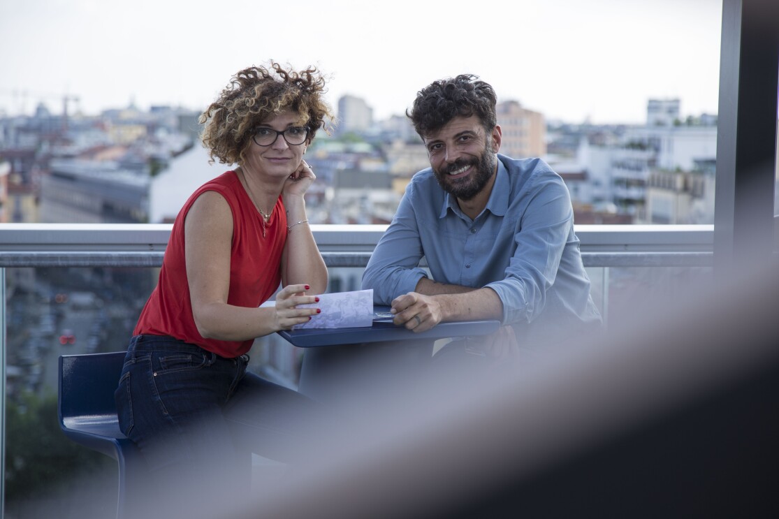 Due persone siedono allo stesso tavolo, sulla terrazza del palazzo Amazon di Milano. A destra, Alessandro Saccon, Consumer PR Manager. Di fronte a lui siede Alessandra Meucci, Executive Assistant. Sullo sfondo lo skyline di Milano. La foto è stata scattata in occasione del Pride di Milano di giugno 2018. 