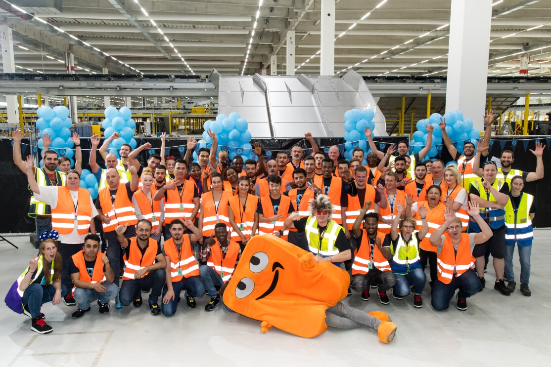 Gruppenbild der Mitarbeiter mit Maskottchen Peccy: Die Mitarbeiter tragen Sicherheitswesten, ein Mitarbeiter trägt das Peccy-Kostüm, das in orangener Farbe an einen Karton erinnert. 