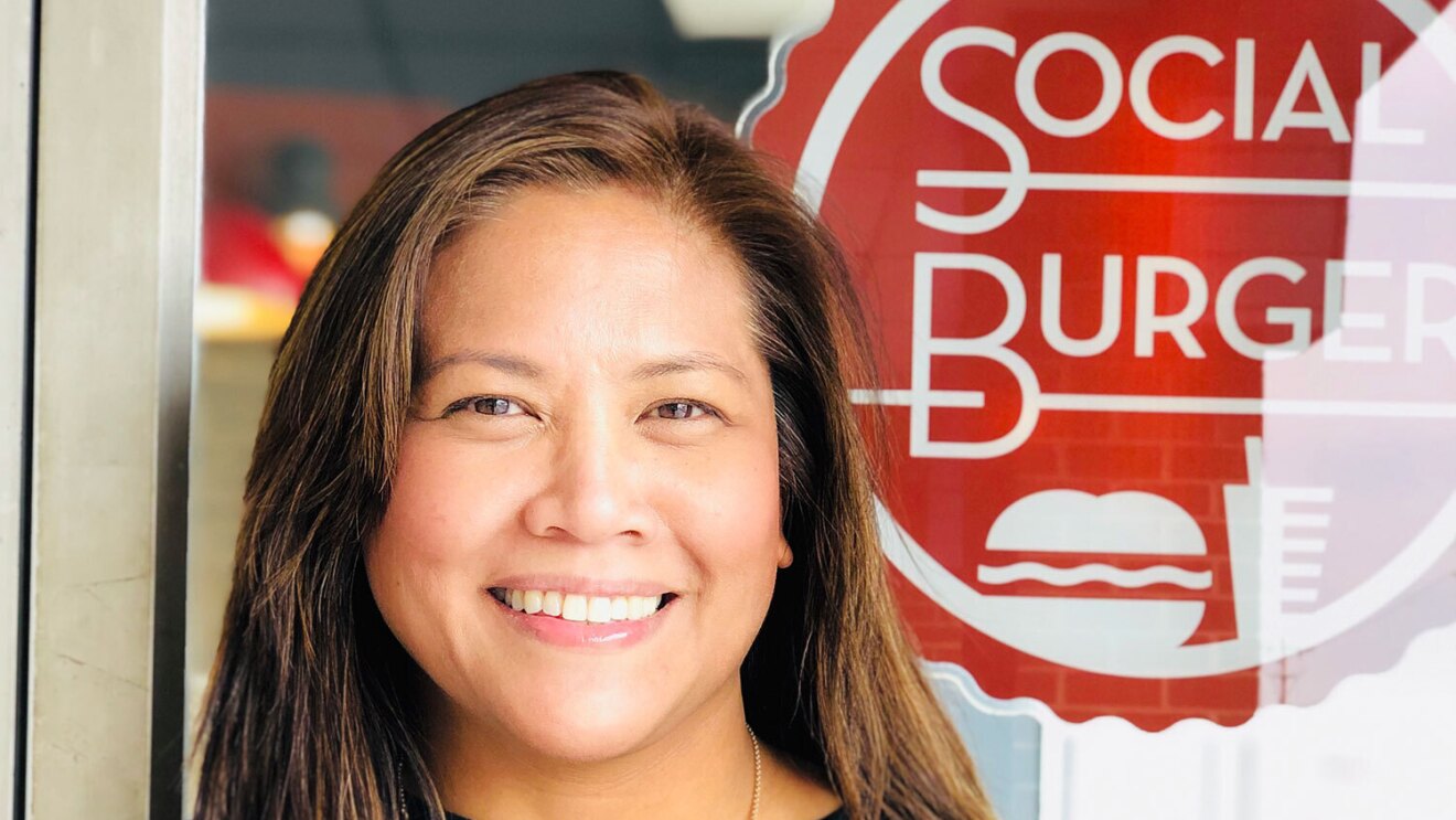 An image of a woman smiling for a photo in front of a window with a red sign that says "Social Burger."