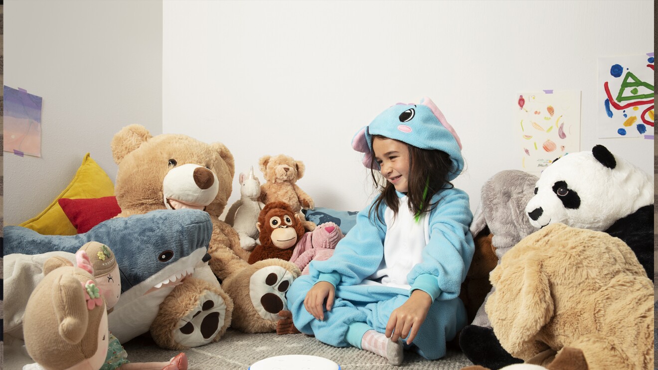 A child sits in a playroom, surrounded by stuffed animals. In front of her is an Echo Dot device is a rainbow stripe, on a tabletop.