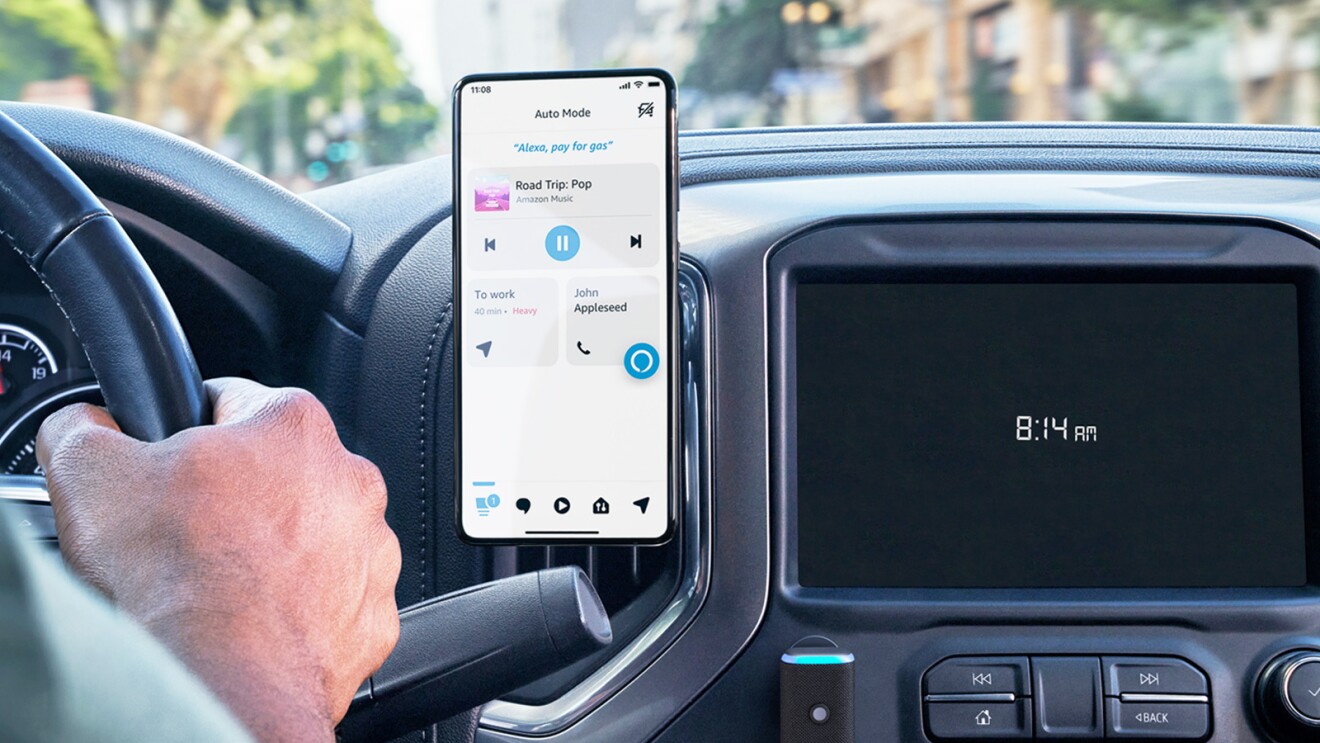 An image of the inside of a car showing part of the steering wheel with a hand on it and the car screen showing 8:14 AM time. There is a phone attached to the car that shows the Alexa Auto Mode setting on the screen. 