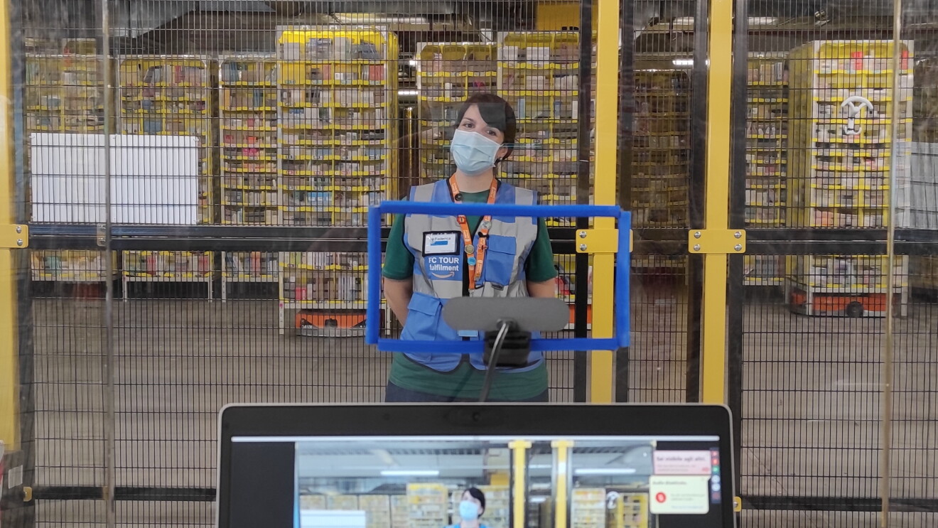 In primo piano un tablet con la fotocamera accesa. Sullo schermo si vede una ragazza all'interno di un magazzino Amazon. La ragazza è posizionata di fronte al tablet, indossa una mascherina chirurgica e un giubbino catarinfrangente blu con la scritta FC Tour Ambassador. Alle sue spalle, una griglia la separa dall'area robotica del magazzino, in cui si vedono le colonne di prodotti trasportate dai robottini. 