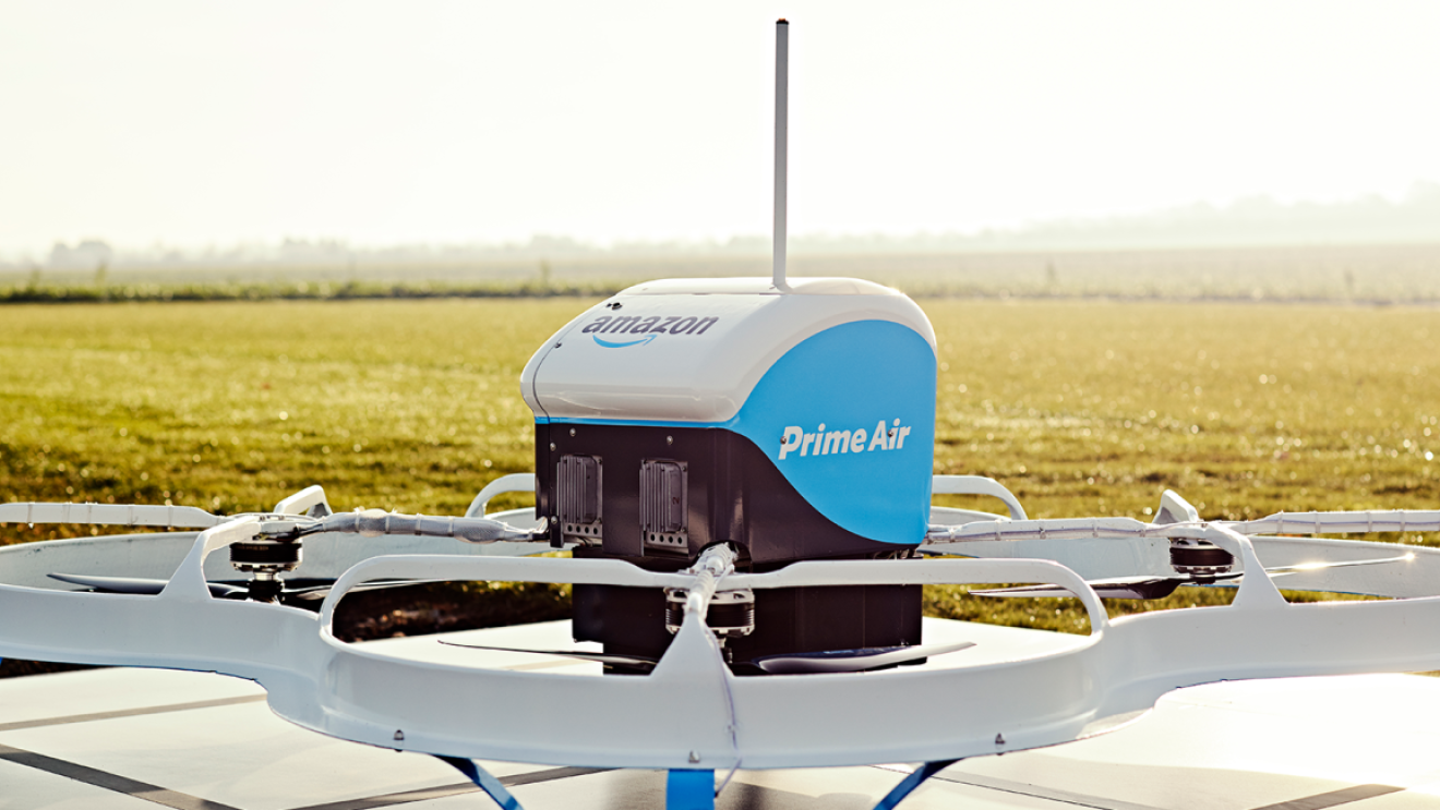 Biało-niebieski dron z logiem Prime Air oraz Amazon, umiejscowiony na metalowej powierzchni, w tle szerokie pole