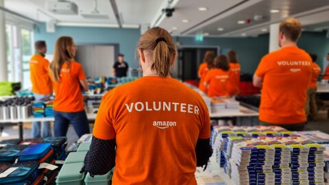 Man sieht Amazon Mitarbeiter:innen mit orangenem Volunteer-T-Shirt von hinten. Sie stehen hinter Tischen mit Schulmaterialien.