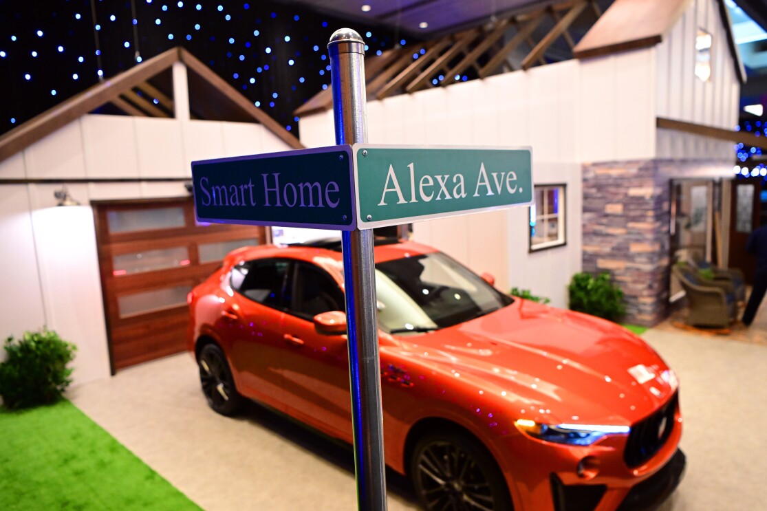 An image of a drive way with an orange car parked on it. There is a street sign in the foreground that says "Smart Home" on one street and "Alexa Ave." on the other.