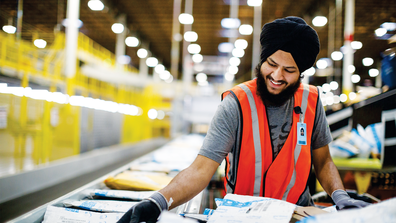 A man, wearing a dastaar (or turban) in an Amazon fulfilment center (warehouse). He is moving packages from a conveyor belt to a large cart to be shipped. 