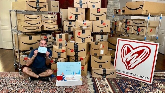 亚马逊的盒子堆在一起,一名男子坐在盒子前,手里拿着为今年活动选定的慈善机构发出的“传递微笑”邀请。还有一个标志,上面有一个大大的心形和“志愿者中心”。"width=