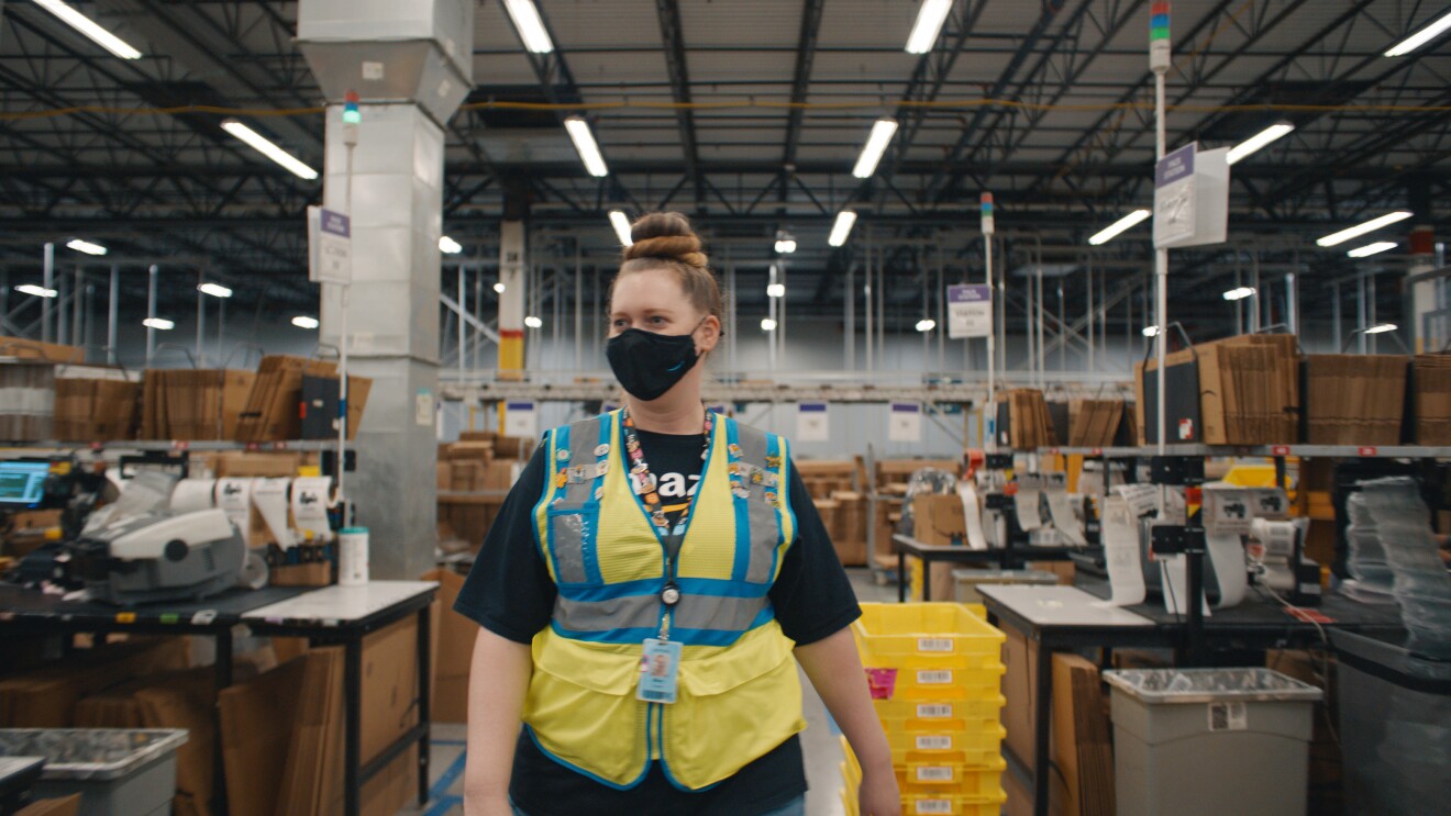 An image of a woman walking through a walkway at Amazon. She is wearing a black mask with the Amazon logo on it and a yellow safety vest. She also had a lanyard with her employee badge on it and the lanyard has many Amazon-related pins on it.