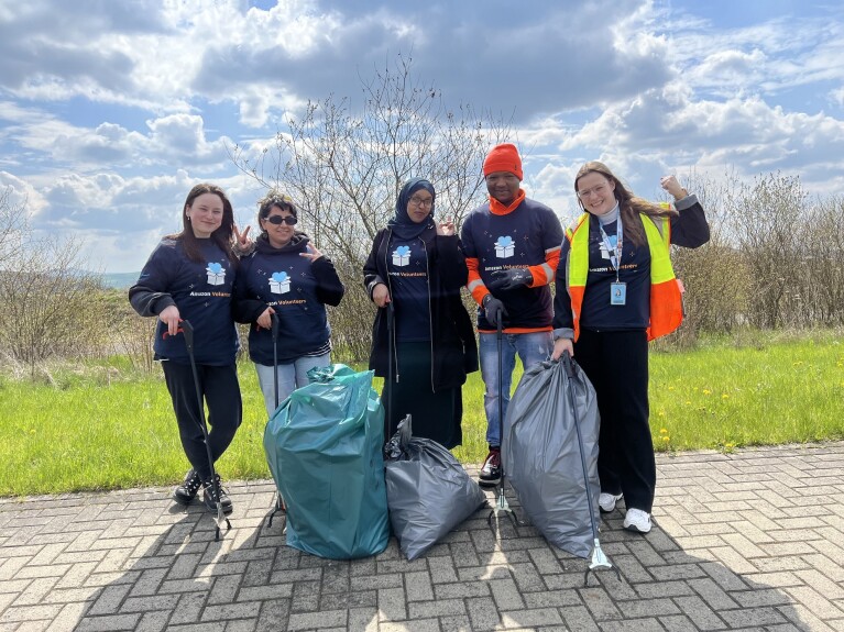 Gruppenfoto der Mitarbeiter:innen vom Amazon Sortierzentrum Breuna/Wolfhagen auf dem Gehweg vor einer grünen Wiese und blauem Himmel.