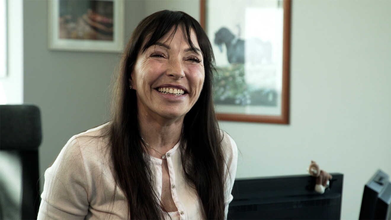 Primo piano di Laura, dipendente Amazon protagonista della storia. La donna ha i capelli mori, ride verso la camera. Alla sue spalle alcuni monitor e un quadro appeso alla parete