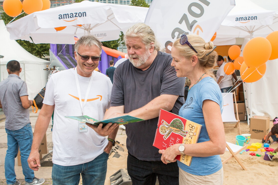 Rulo Lange ist in der Bildmitte zu sehen. Links Standortleiter Dietmar Jüngling, rechts eine Dame. Rulo Lange zeigt beiden ein KInderbuch. Im Hintergrund ist der SachsenBeach in Leipzig zu sehen, der von Amazon gesponsert wurde.