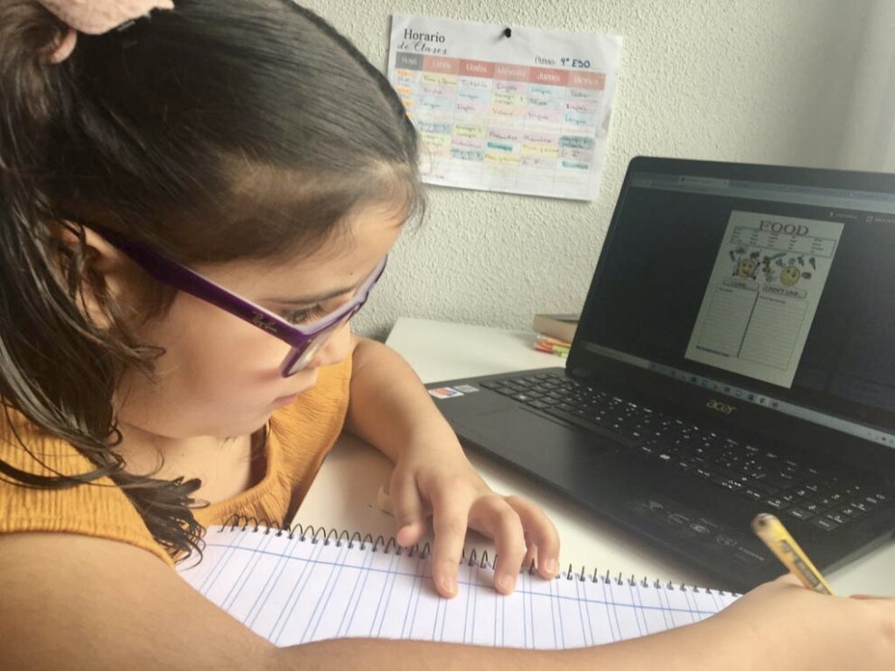 Hay una niña de 4 de ESO con gafas moradas, pelo rizado por el hombro y con un lápiz en la mano y una libreta. Está en su escritorio con un ordenador que hay una página de deberes con la palabra inglesa food. De fondo una pared blanco y un horario escolar colgado. 