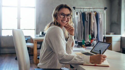 Una donna sorridente seduta nel suo ufficio mentre annota qualcosa su una agenda. Indossa degli occhiali da vista. Sullo sfondo, una rella con degli abiti appesi