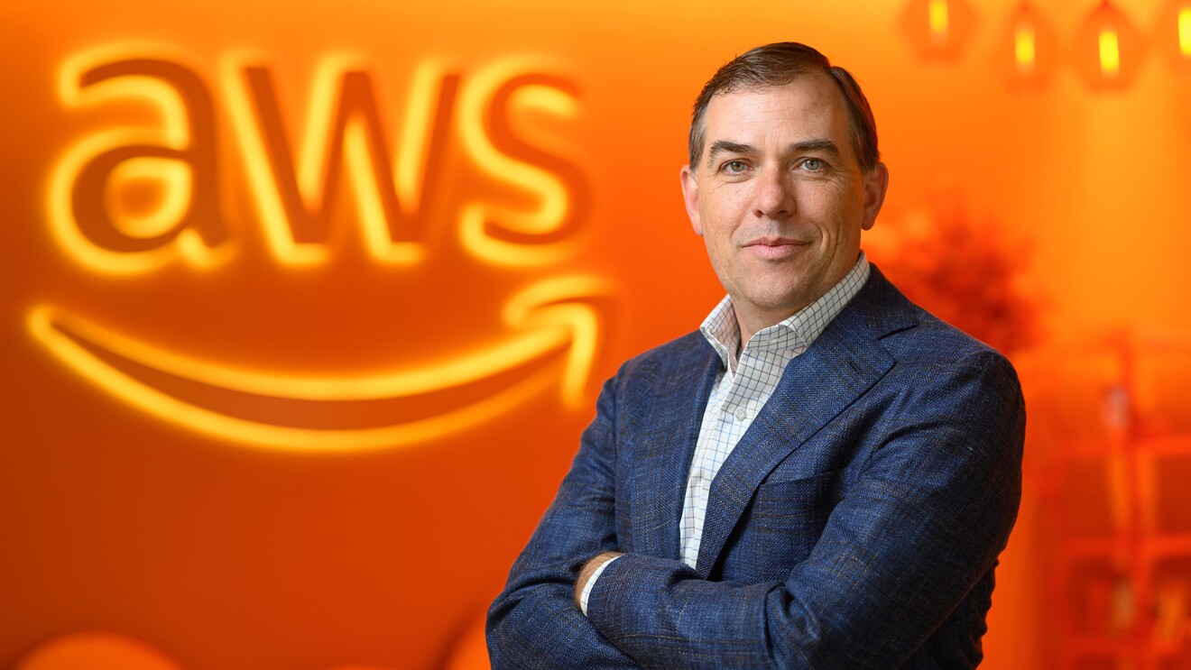 A photo of AWS CEO Matt Garman with his arms crossed. Garman is standing in front of a wall that has a lit sign of the AWS logo.