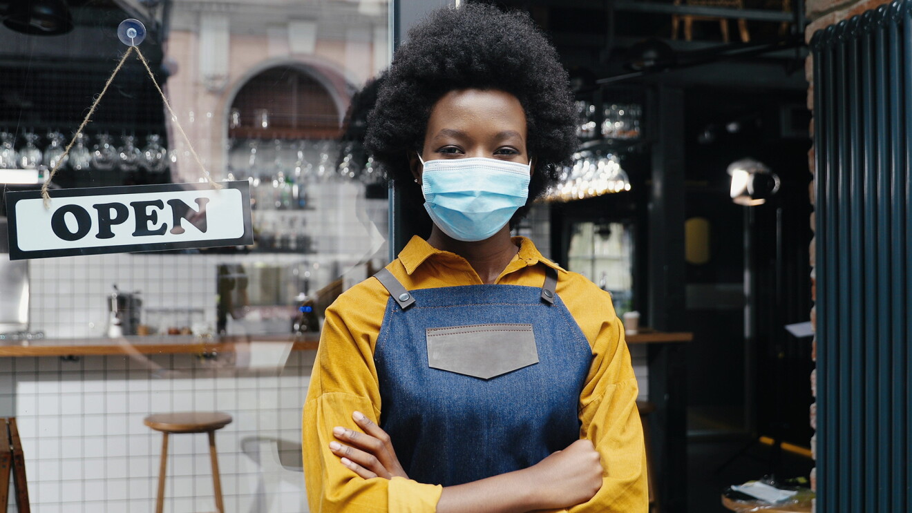 A Black woman wearing a mask and a denim apron stands in the open doorway of a restaurant